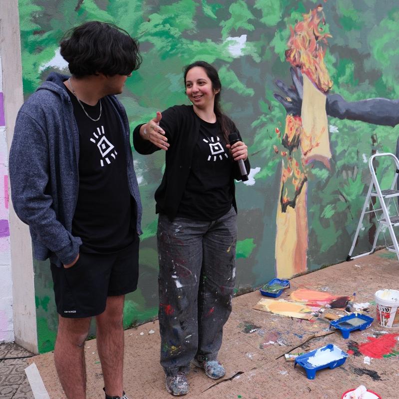 Dues persones parlant davant el mural amb la pintura i estris encara a terra