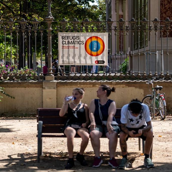 Tres persones assegudes a l'ombra d'un banc. De fons, la cartelleria informativa a un refugi climàtic